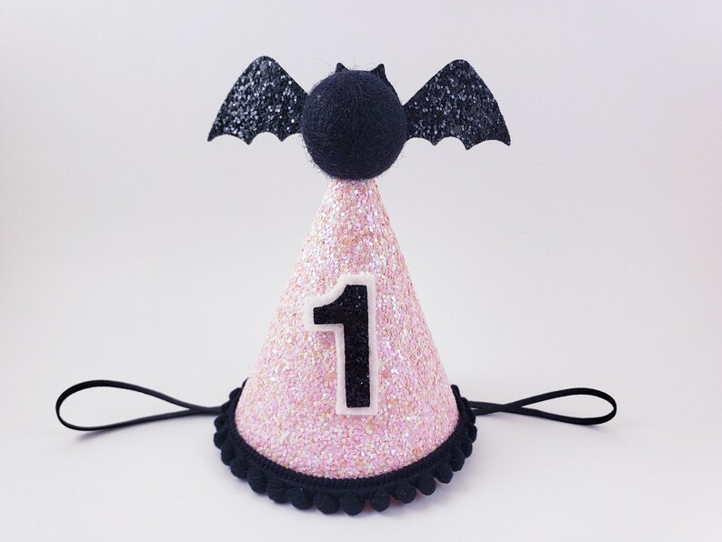 May include: Pink and black glitter party hat with a black bat on top and the number "1" on the front. The hat has a black elastic band.