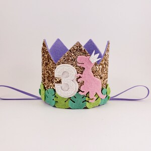 May include: A gold glitter crown with a purple felt top and green felt leaves. A pink felt dinosaur with a white crown is on the front of the crown. The number "3" is on the front of the crown in white felt.