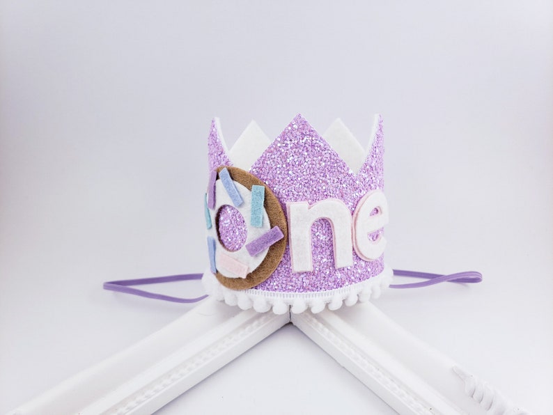May include: Purple glitter crown with a white felt 'one' and a donut with sprinkles. The crown has a purple elastic band.