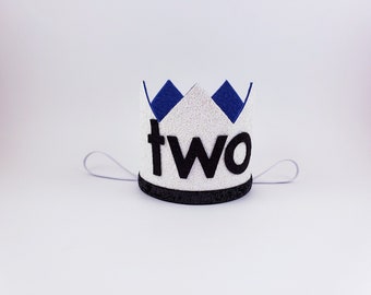 May include: White felt crown with blue felt accents and the word "two" in black felt letters. The crown has a white elastic band.