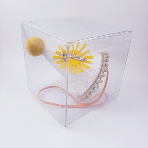 May include: A white party hat with a yellow felt sun and a yellow pom-pom on top. The hat is in a clear plastic box with a brown ribbon.