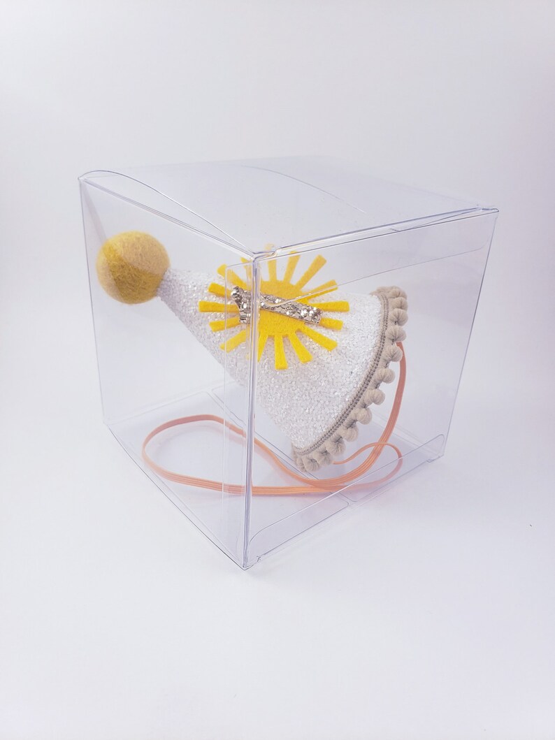 May include: A white party hat with a yellow felt sun and a yellow pom-pom on top. The hat is in a clear plastic box with a brown ribbon.