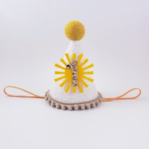 May include: White felt party hat with a yellow sun and the number one in gold glitter. The hat has a tan pom-pom trim and an orange elastic band.