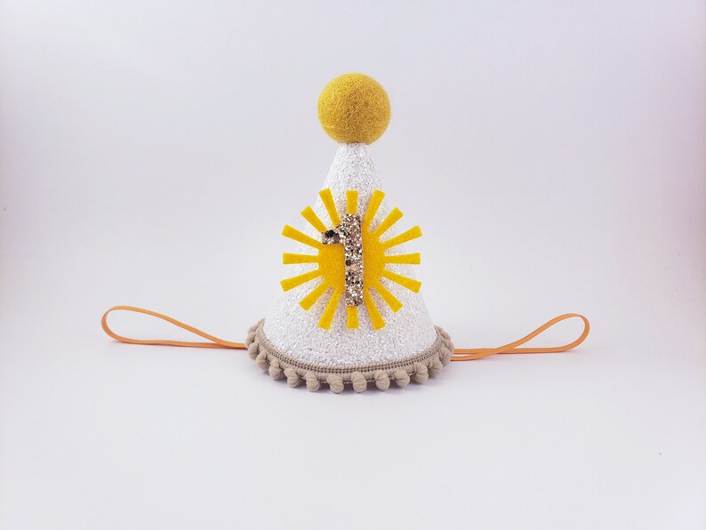 May include: White felt party hat with a yellow sun and the number one in gold glitter. The hat has a tan pom-pom trim and an orange elastic band.