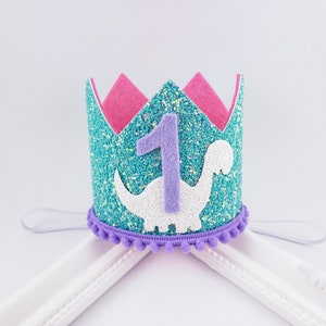 May include: A turquoise glitter crown with a purple felt number one and a white felt dinosaur. The crown has a purple felt band with pom poms around the edge.