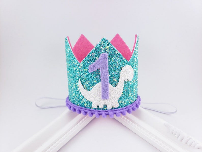 May include: A turquoise glitter crown with a purple felt number one and a white felt dinosaur. The crown has a purple felt band with pom poms around the edge.