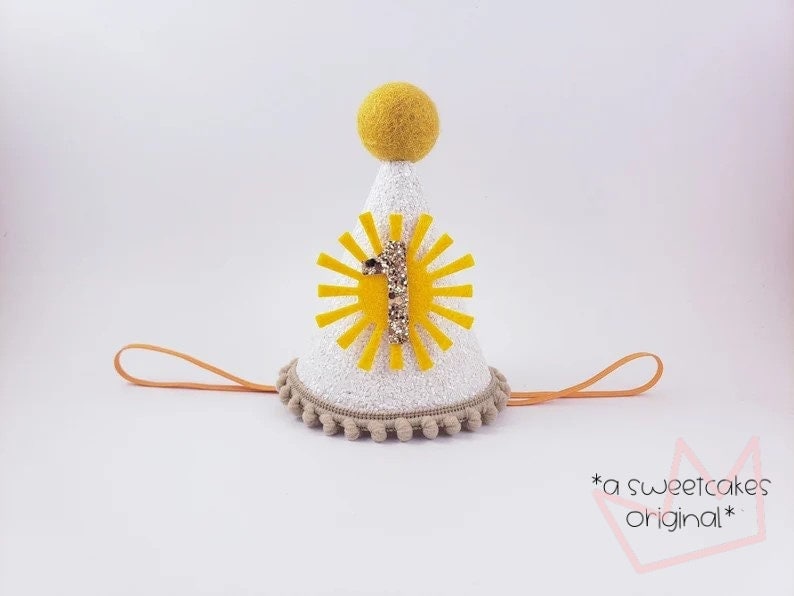 May include: White felt party hat with a yellow sun and the number one on it. The hat has a brown pom-pom trim and a brown elastic band.