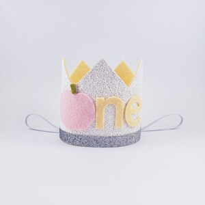 May include: A white felt crown with silver glitter, a pink felt peach, and the word "one" in yellow felt. The crown has a white elastic band.
