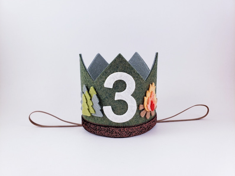 May include: A green felt crown with a white felt number "3" in the center. The crown is decorated with felt trees and a campfire. The crown has a brown felt tie.