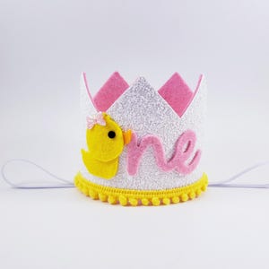 May include: White felt crown with pink felt accents and a yellow felt duck. The crown has a white glitter finish and says "one" in pink felt. The crown has a yellow pom-pom trim.