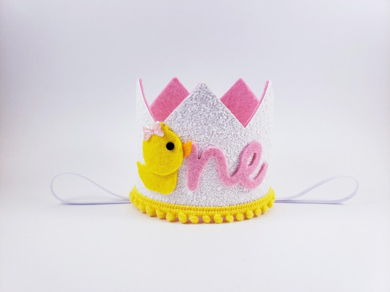May include: White felt crown with pink felt accents and a yellow felt duck. The crown has a white glitter finish and says "one" in pink felt. The crown has a yellow pom-pom trim.