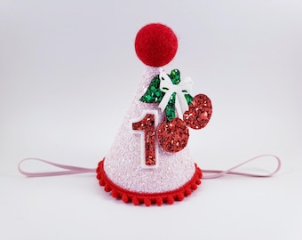 May include: A pink glitter party hat with a red pom-pom, a red pom-pom trim, and the number 1. The hat is decorated with a white bow, green leaves, and red glitter cherries. A pink elastic band is attached.
