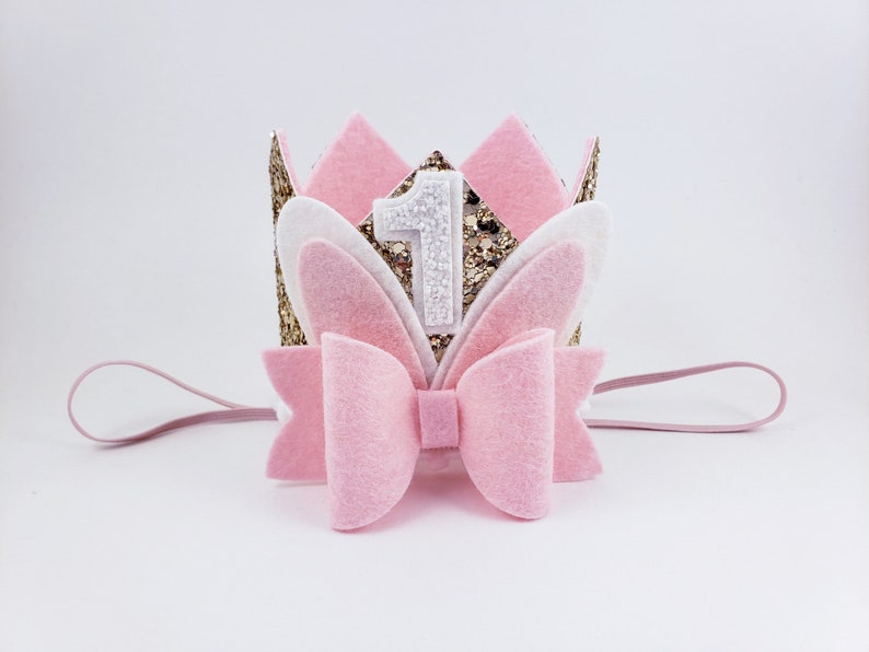 May include: Pink felt headband with a gold glitter crown, white felt bunny ears, and a pink bow. The crown has a white felt number one on it.