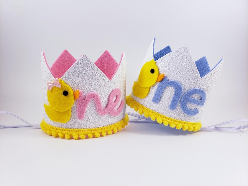 May include: Two white felt crowns with yellow pom-poms around the base. One crown has a pink felt "one" and a yellow felt duck with a pink bow. The other crown has a blue felt "one" and a yellow felt duck. Both crowns have white felt and glitter.