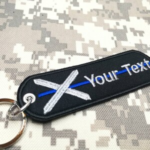 Peut inclure: Porte-clés noir avec un X brodé en blanc et une ligne bleue qui le traverse. Le texte "Your Text" est brodé en blanc sur le porte-clés.