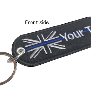 Könnte beinhalten: Schwarzer Schlüsselanhänger mit einem silbernen Union-Jack-Design und einer blauen Linie in der Mitte. Der Text "Your-Text" ist auf den Schlüsselanhänger gestickt.