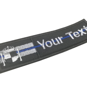 May include: Black embroidered patch with a thin blue line and a white and gray emblem of a crown and shield. The text "Your-Text" is embroidered in white.