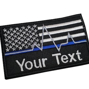 May include: Black and white embroidered patch with an American flag, a blue line, and a heartbeat graphic. The text "Your Text" is embroidered below the graphic.