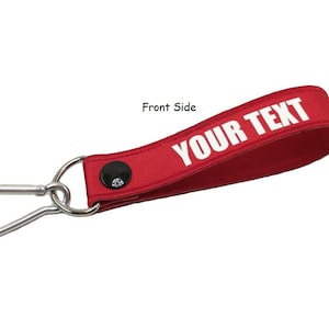 Puede incluir: Llavero de tela roja con un mosquetón plateado. La tela tiene el texto "YOUR TEXT" impreso en blanco.