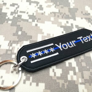 May include: Black and blue keychain with a thin blue line and five white stars. The keychain has a silver ring and the text "Your-Text" is embroidered on the black fabric.