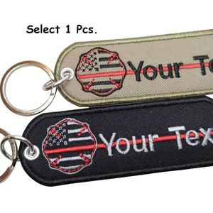 May include: Two keychains with a red line and a thin blue line American flag design. The keychains are available in black and tan with the text "Your Text" embroidered on them.
