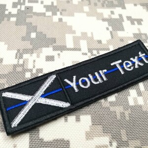May include: Black and white embroidered patch with a blue line across the center. The patch has a white cross on a black background and the text "Your Text" in white.