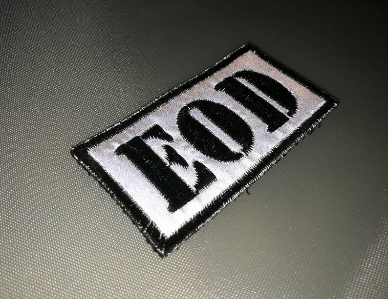 K&ouml;nnte beinhalten: Ein schwarz-wei&szlig;es Patch mit dem Wort "EOD" in Schwarz auf wei&szlig;em Hintergrund gestickt. Das Patch hat einen schwarzen Rand und ist auf einer grauen Oberfl&auml;che aufgen&auml;ht.