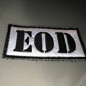 K&ouml;nnte beinhalten: Ein gestickter schwarzer und wei&szlig;er Aufn&auml;her mit dem Wort "EOD" in wei&szlig;en Buchstaben auf schwarzem Hintergrund.