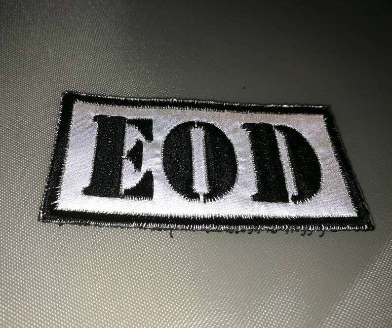 K&ouml;nnte beinhalten: Ein gestickter schwarzer und wei&szlig;er Aufn&auml;her mit dem Wort "EOD" in wei&szlig;en Buchstaben auf schwarzem Hintergrund.