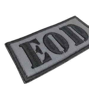 K&ouml;nnte beinhalten: Ein grauer und schwarzer gestickter Aufn&auml;her mit den Buchstaben "EOD" in Schwarz.