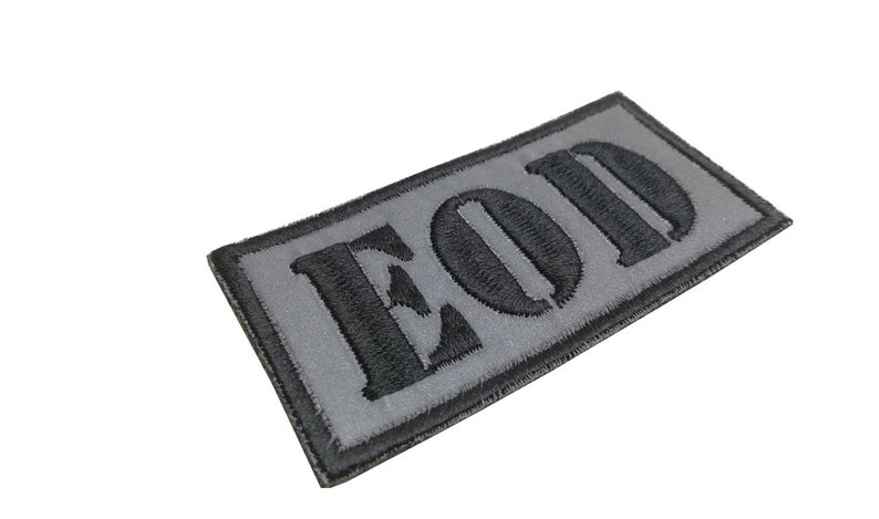 K&ouml;nnte beinhalten: Ein grauer und schwarzer gestickter Aufn&auml;her mit den Buchstaben "EOD" in Schwarz.