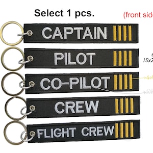 Może przedstawiać: Pięć czarnych breloków z różnymi białymi etykietami tekstowymi: "CAPTAIN", "PILOT", "CO-PILOT", "CREW" i "FLIGHT CREW". Każdy brelok ma złoty pasek i srebrny kółko do kluczy.
