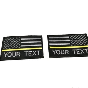Op de afbeelding: Twee zwarte geborduurde patches met een dunne gele streep aan de onderkant. De patches tonen een zilveren geborduurde Amerikaanse vlag met de tekst "YOUR TEXT" eronder.