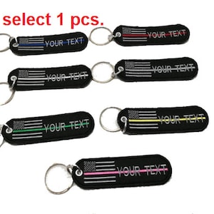 Op de afbeelding: Vijf verschillende sleutelhangers met een Amerikaans vlagontwerp en een dunne gekleurde lijn over de vlag. Elke sleutelhanger heeft de tekst "YOUR TEXT" erop gedrukt. De gekleurde lijnen zijn rood, blauw, zilver, groen en roze.