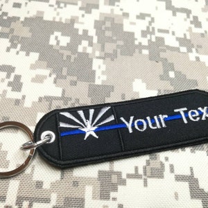 May include: Black rectangular keychain with a silver key ring. The keychain features the Arizona state flag design with a blue line and the text "Your Text" in white.