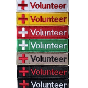 Peut inclure: Plusieurs écussons rectangulaires brodés de différentes couleurs, dont blanc, jaune, rouge, vert, beige, noir et gris foncé. Chaque écusson comporte une croix rouge et le mot "Volunteer". Dimensions approximatives : 10 cm de long x 2,5 cm de haut.
