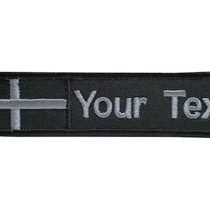 May include: Black rectangular patch with gray embroidered text that reads "Your Text." A gray cross design is on the left side. The patch is likely for clothing or accessories.