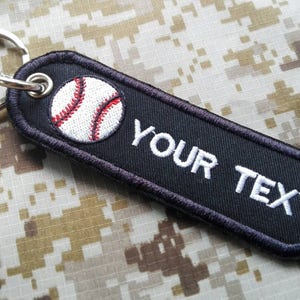 May include: Black key chain with a white baseball and the text "YOUR TEXT" embroidered on it.