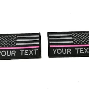 Pode incluir: Dois patches de bandeira americana em preto e prata com uma faixa rosa na parte inferior. O texto "YOUR TEXT" é bordado em branco abaixo da faixa.