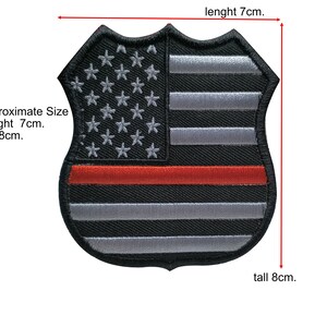 Può includere: Un patch di bandiera americana in bianco e nero con una striscia rossa al centro. Il patch ha la forma di uno scudo e misura circa 7 cm di lunghezza e 8 cm di altezza.