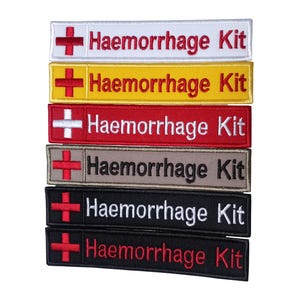 Może przedstawiać: Sześć haftowanych naszywek z czerwonym krzyżem i napisem "Haemorrhage Kit" w kolorze białym. Naszywki są w różnych kolorach: białym, żółtym, czerwonym, szarym, czarnym i czarnym z czerwonym napisem.