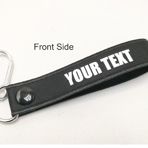 Könnte beinhalten: Schwarzer Schlüsselanhänger mit einem silbernen Metallhaken und einem schwarzen Band mit weißem Text, der "YOUR TEXT" lautet.