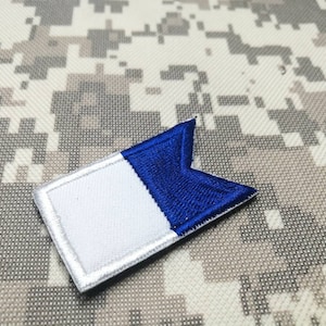 Puede incluir: Un parche rectangular con un diseño blanco y azul. El parche está dividido en dos secciones, con el lado izquierdo blanco y el lado derecho azul. Tiene un borde blanco y está sobre un fondo de camuflaje.