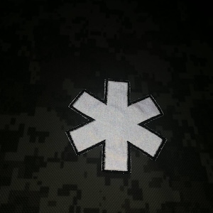 Könnte beinhalten: Ein weißer reflektierender Stern-des-Lebens-Patch auf einem dunklen Tarnmuster-Hintergrund.