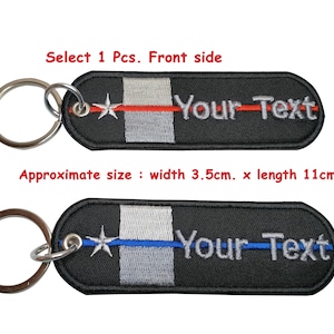 Puede incluir: Dos llaveros negros con un anillo de llave plateado. Los llaveros están bordados con una estrella blanca, una fina línea roja o azul y el texto "Your Text". Los llaveros miden aproximadamente 3,5 cm de ancho y 11 cm de largo.