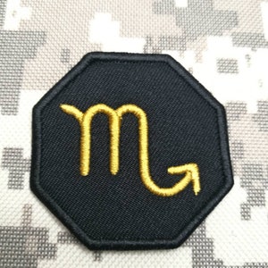 May include: Black embroidered patch with a gold scorpion symbol in the center. The patch is shaped like an octagon.