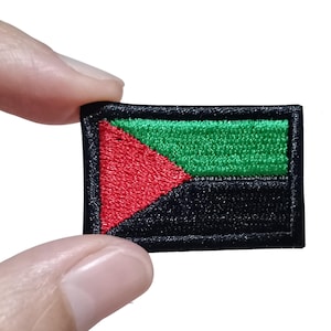 May include: A rectangular embroidered patch featuring a design with a red triangle on the left side, a green horizontal stripe on the top right, and a black horizontal stripe on the bottom right, all bordered by a thick black stitched edge.