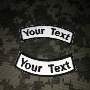 Peut inclure: Deux patchs brodés blancs avec des contours noirs, en forme de bannières courbes. Le texte "Your Text" est sur chaque patch.