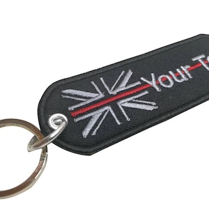 Könnte beinhalten: Schwarzer Schlüsselanhänger mit einem silbernen Union-Jack-Design und einem roten Streifen in der Mitte. Der Schlüsselanhänger hat einen silbernen Ring und der Text "Your-Text" ist in Weiß gestickt.