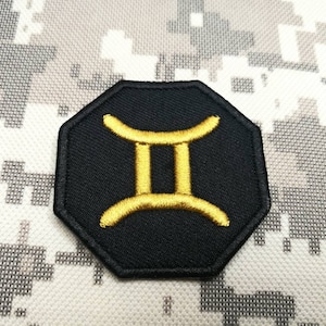 Puede incluir: Un parche octogonal negro con un símbolo zodiacal de Géminis bordado en oro.